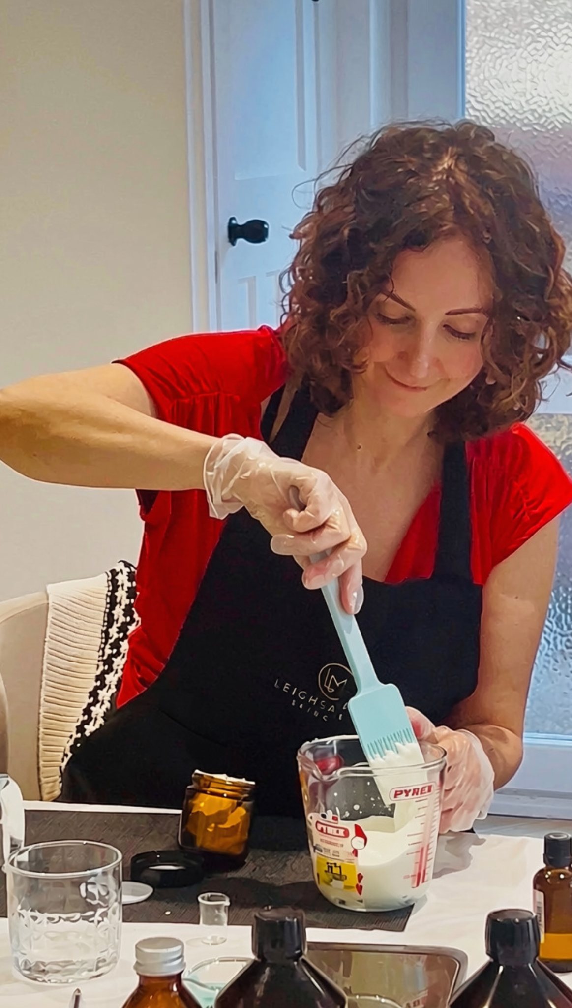 Woman in black apron with gold Leighsa Mari logo, using spatula to scoop body butter mixture in jar