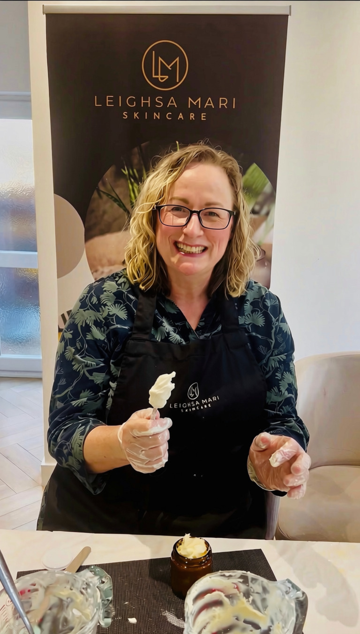 Woman wearing black apron with gold Leighsa Mari logo, with spoon in and filling jar with body butter mixture